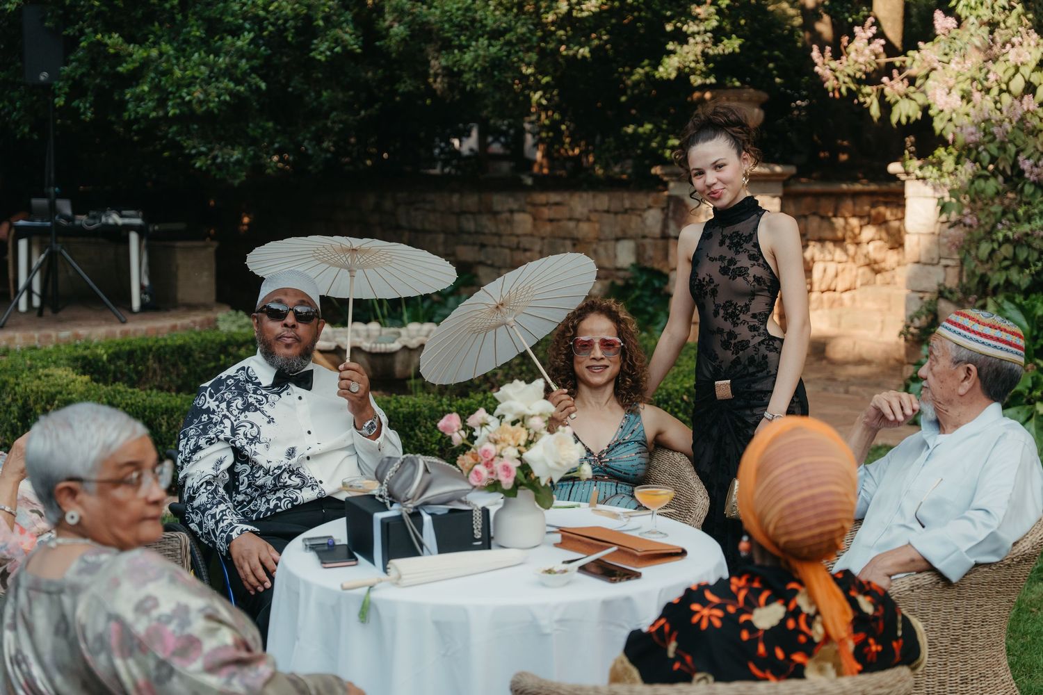 Guests celebrate outdoors at a garden wedding reception, holding parasols and flowers at a decorated table.