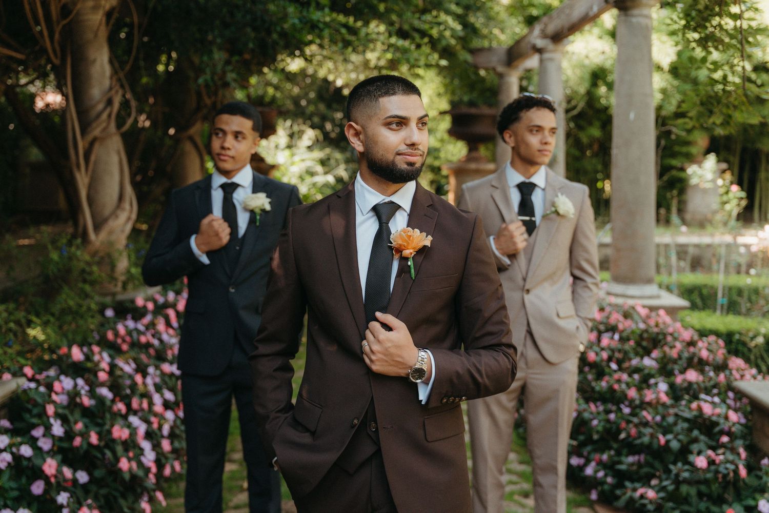 Groom in brown suit with boutonniere stands confidently outdoors, flanked by two groomsmen in navy and tan suits.