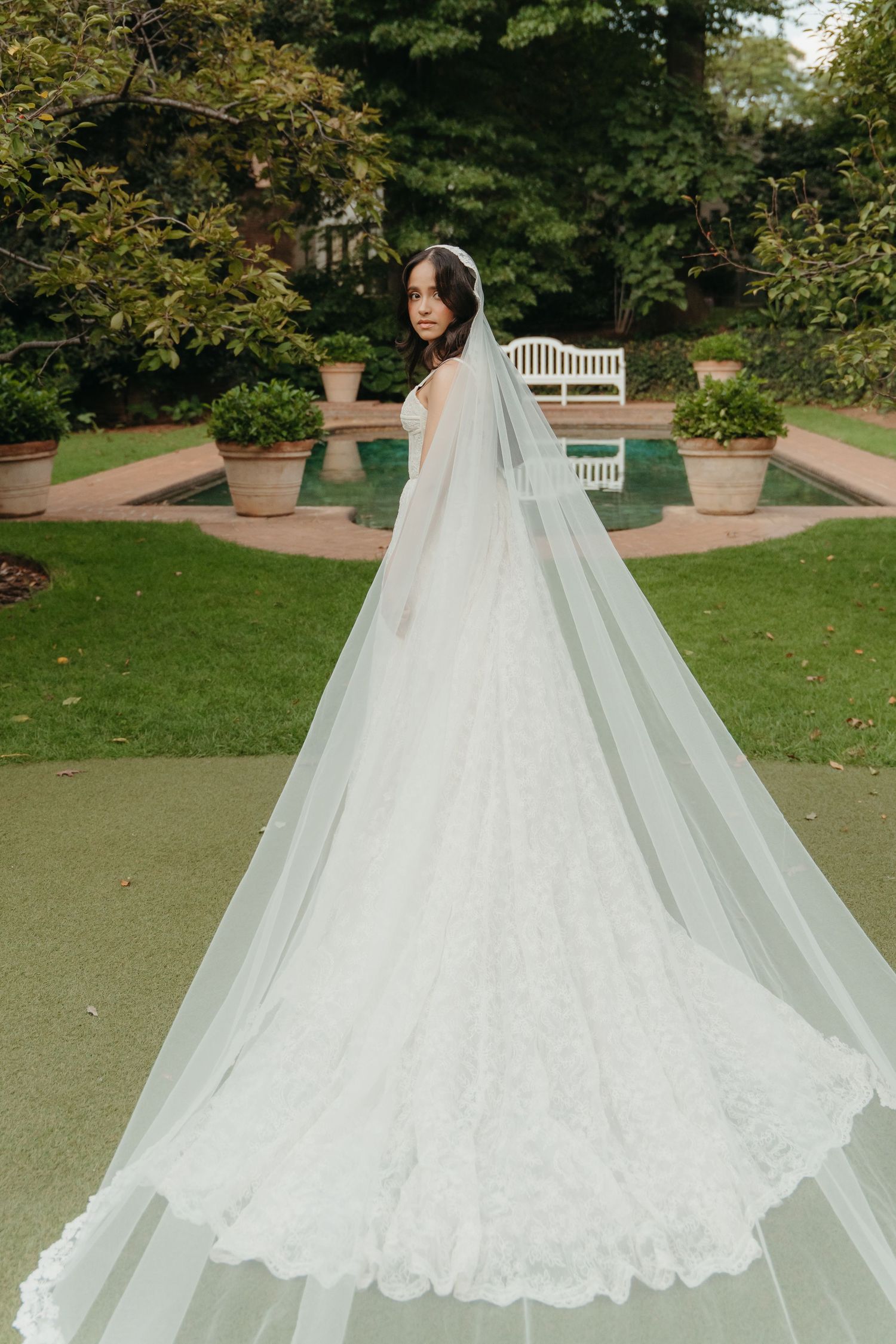 Bride in a stunning lace wedding gown with a long cathedral veil, posing in an elegant garden setting.