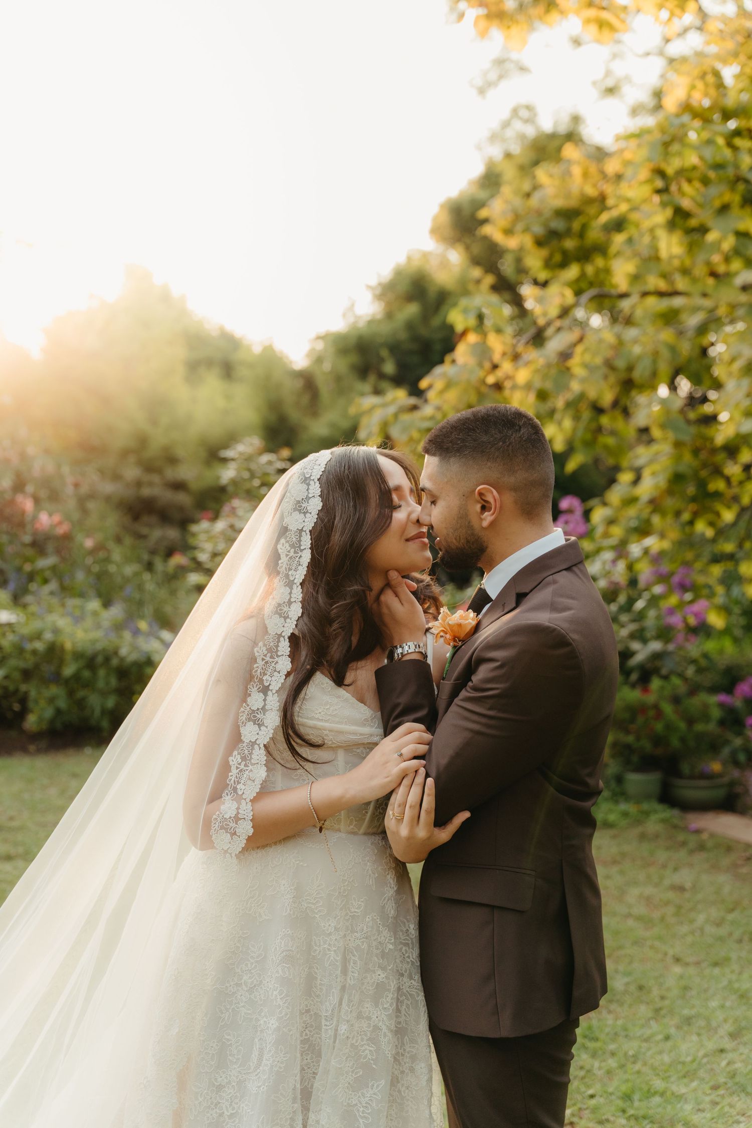 Bride and groom share a romantic moment in a sunlit garden, with bride wearing a lace veil and groom in a brown suit.