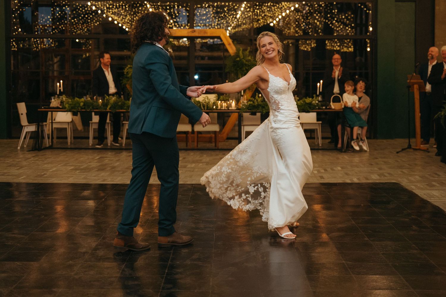 Bride and groom share first dance at elegant indoor wedding reception with warm string lights and guests seated nearby.