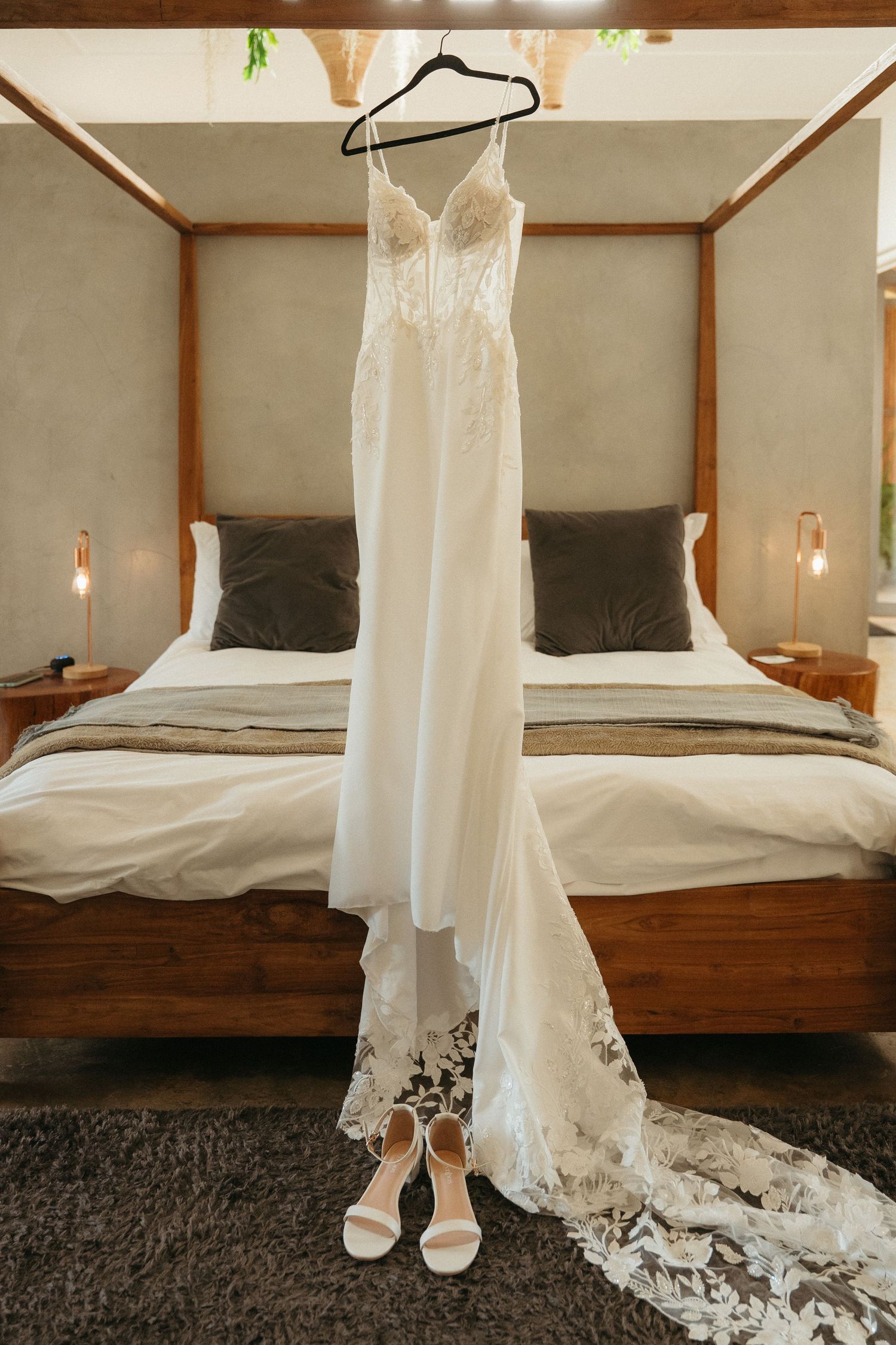 White lace wedding dress hanging on a four-poster bed frame with matching bridal heels displayed below.