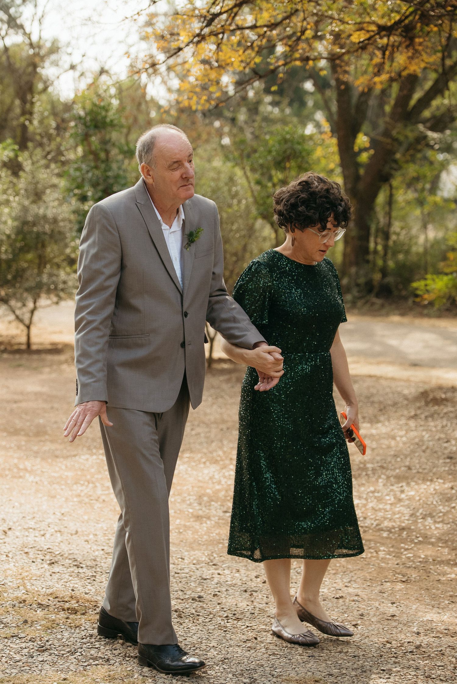 Elderly couple walking hand in hand on a gravel path, man in gray suit, woman in green dress, outdoor setting.