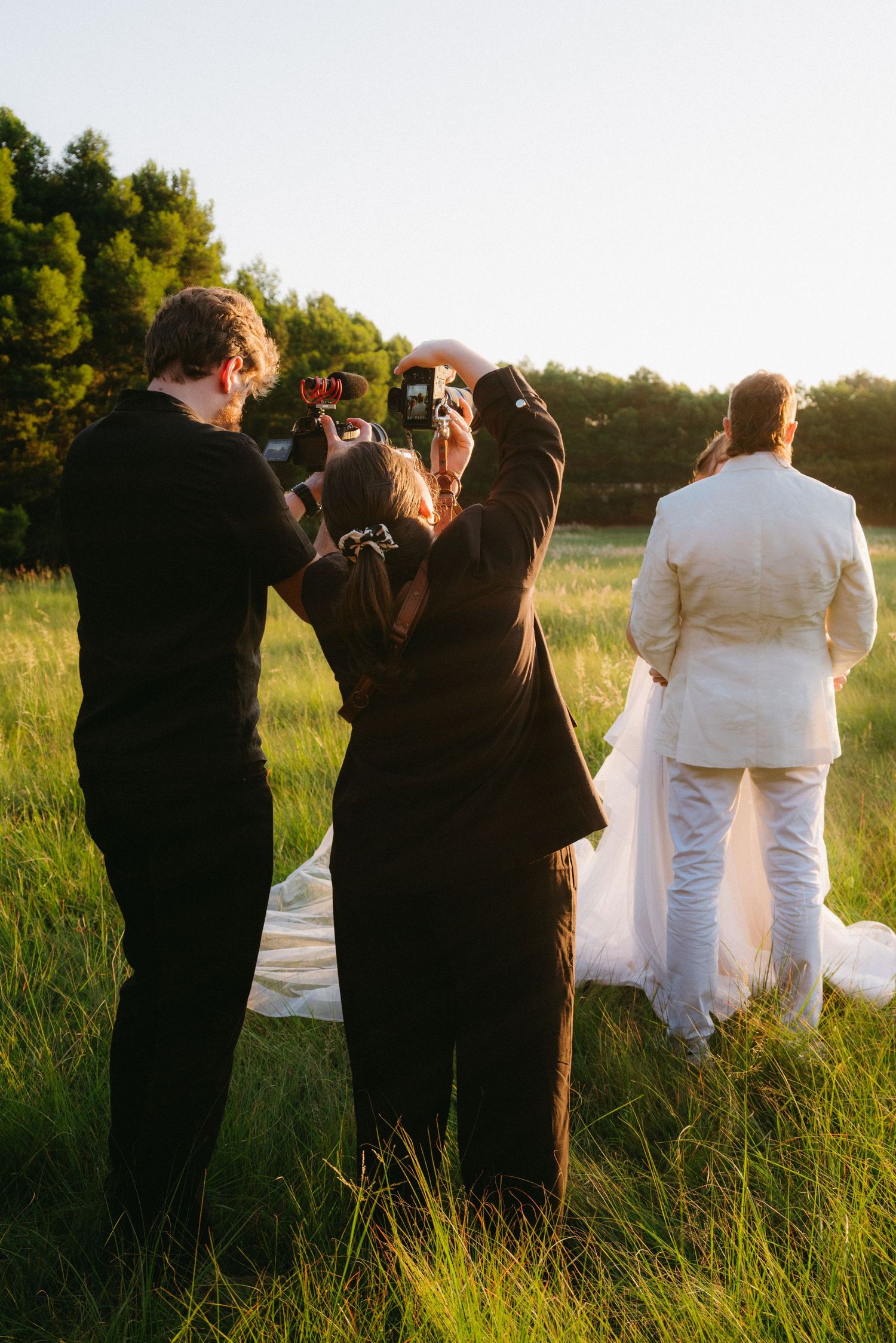 Photographer and videographer capturing a bride and groom from behind in a sunlit green field at golden hour in Pretoria