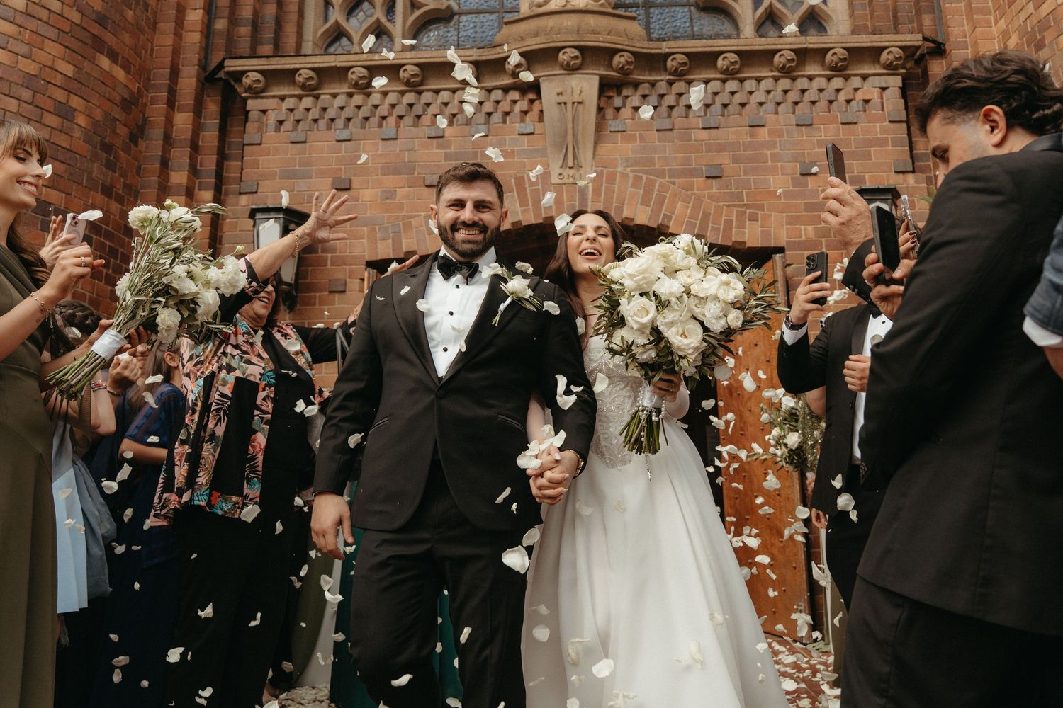 Newlywed couple walks through confetti shower outside Rosebank Catholic Church, Johannesburg with bride holding white bouquet