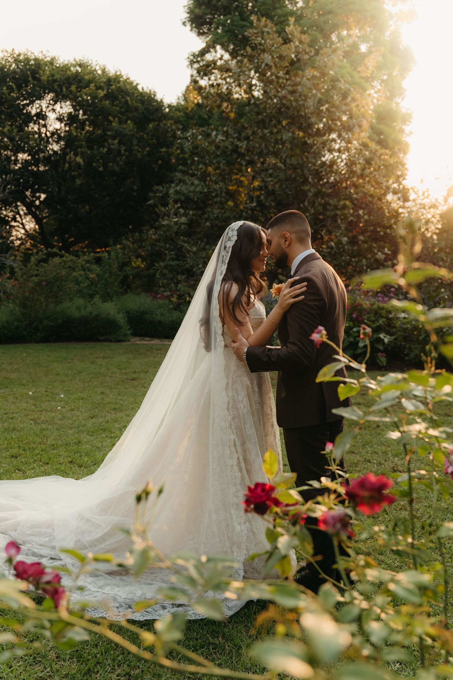Bride and groom share intimate moment at Beechwood Gardens at golden hour, wedding veil trailing on grass.