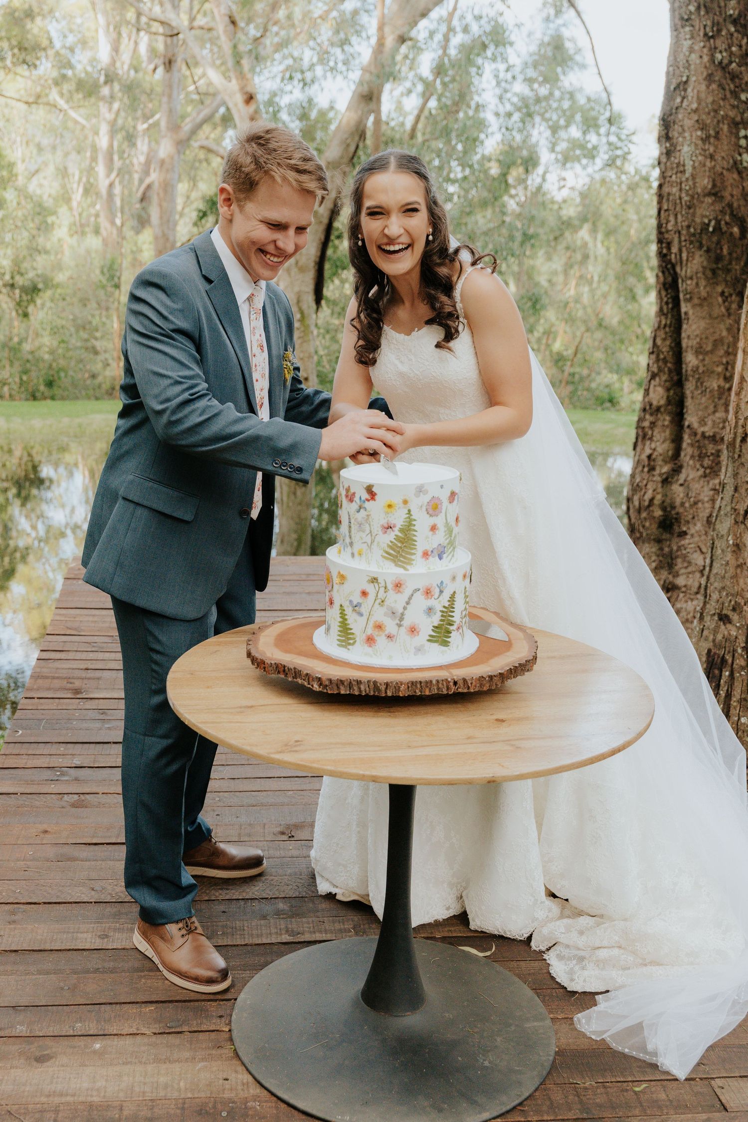 A couple cuts their floral wedding cake together on a wooden deck surrounded by trees in a romantic outdoor setting.