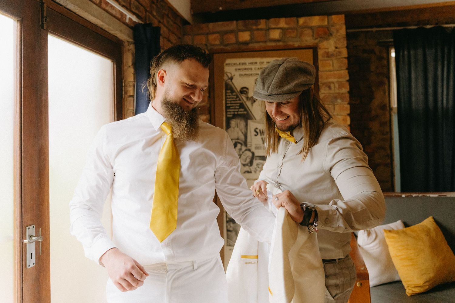 Groom and companion sharing a relaxed moment indoors while wearing white and yellow wedding attire.