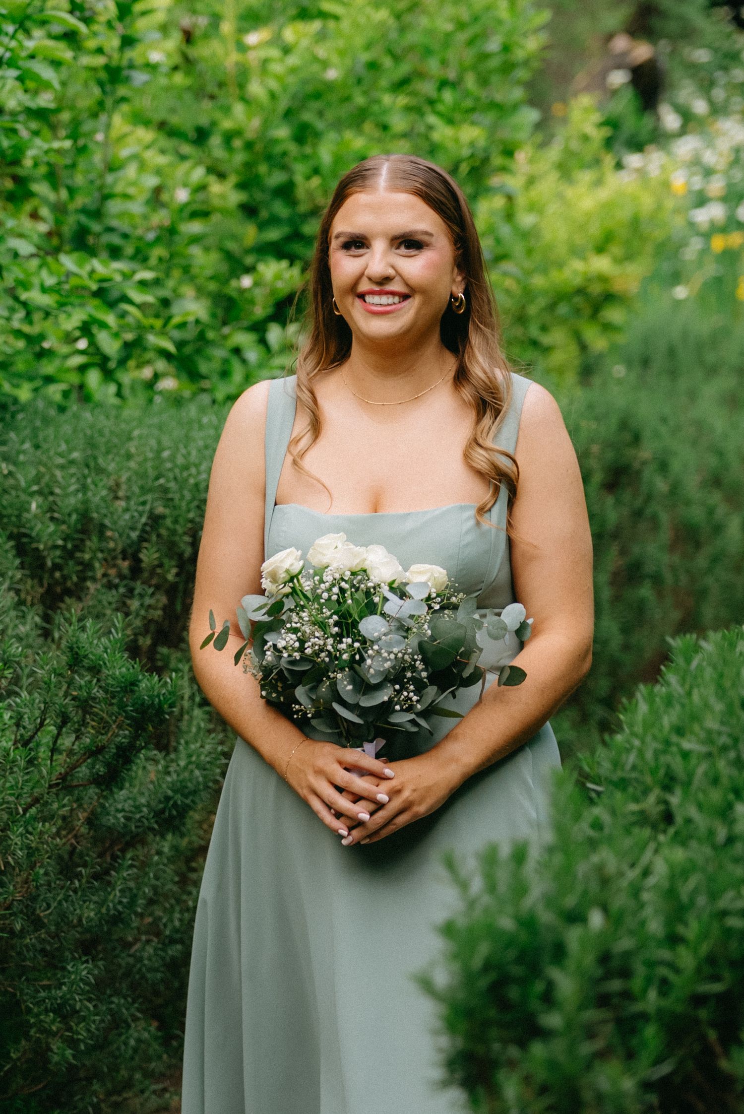 Bridesmaid in a sage green dress holds a delicate bouquet against a lush garden backdrop.