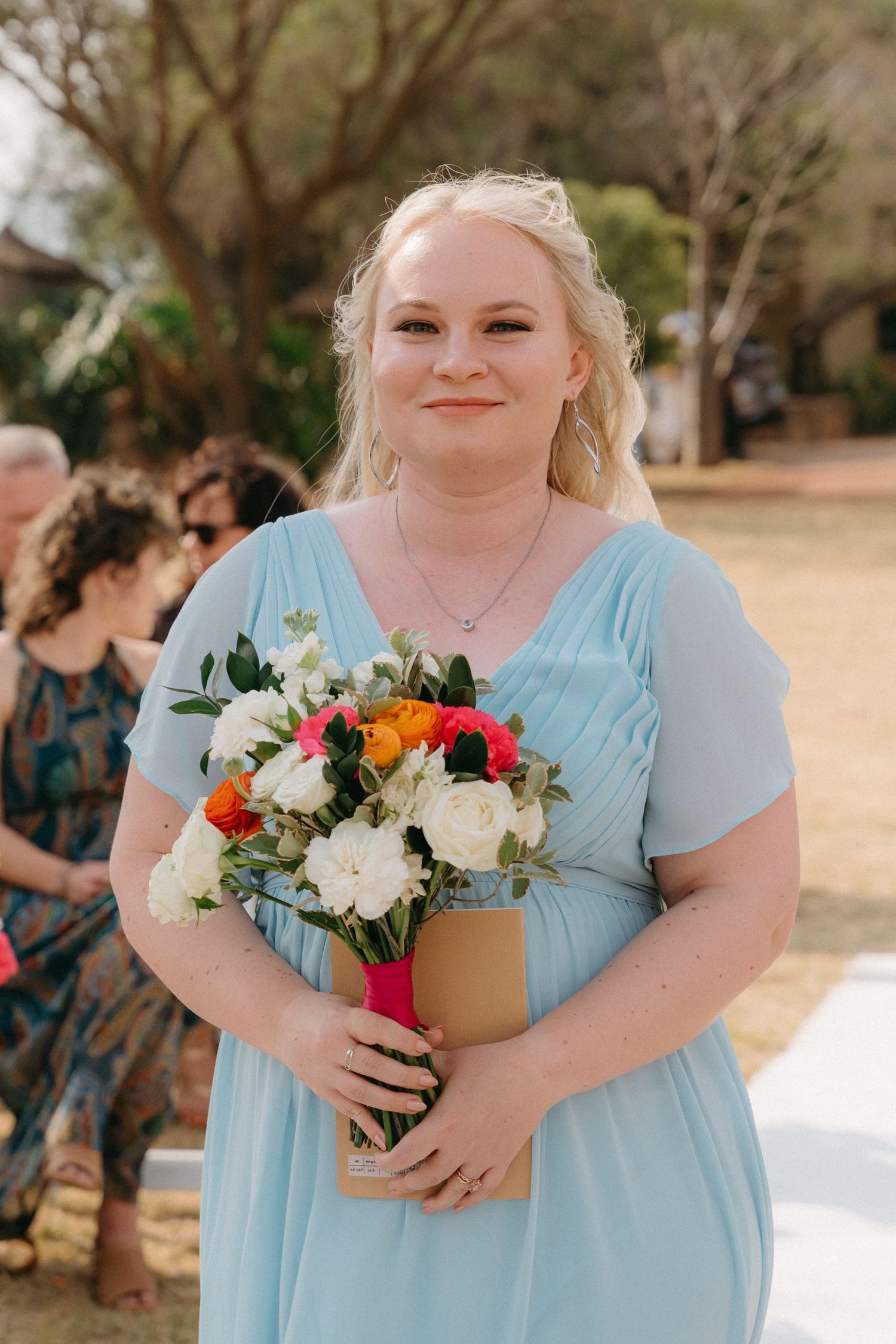 Bridesmaids in light blue dresses hold colorful bouquets of white and red flowers at an outdoor wedding ceremony.