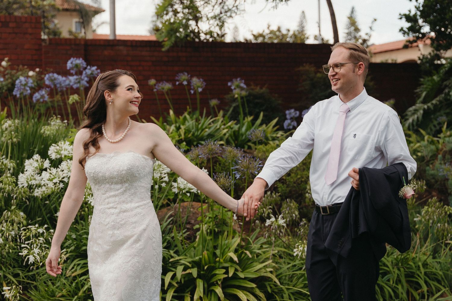 A couple in wedding attire dances joyfully together in a lush garden setting with greenery and flowers.