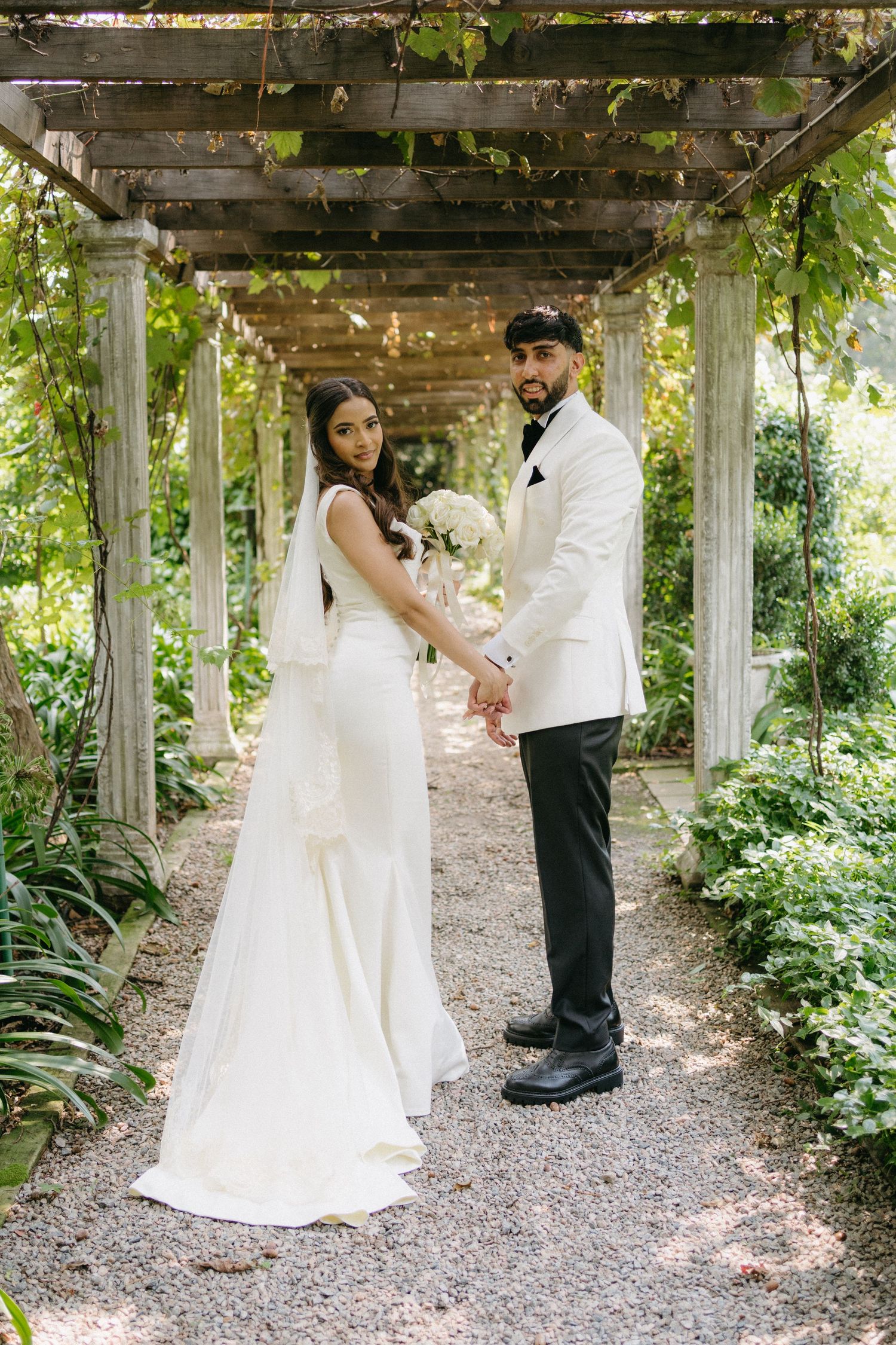 Newlyweds pose under a vine-covered garden pergola in elegant white wedding attire.