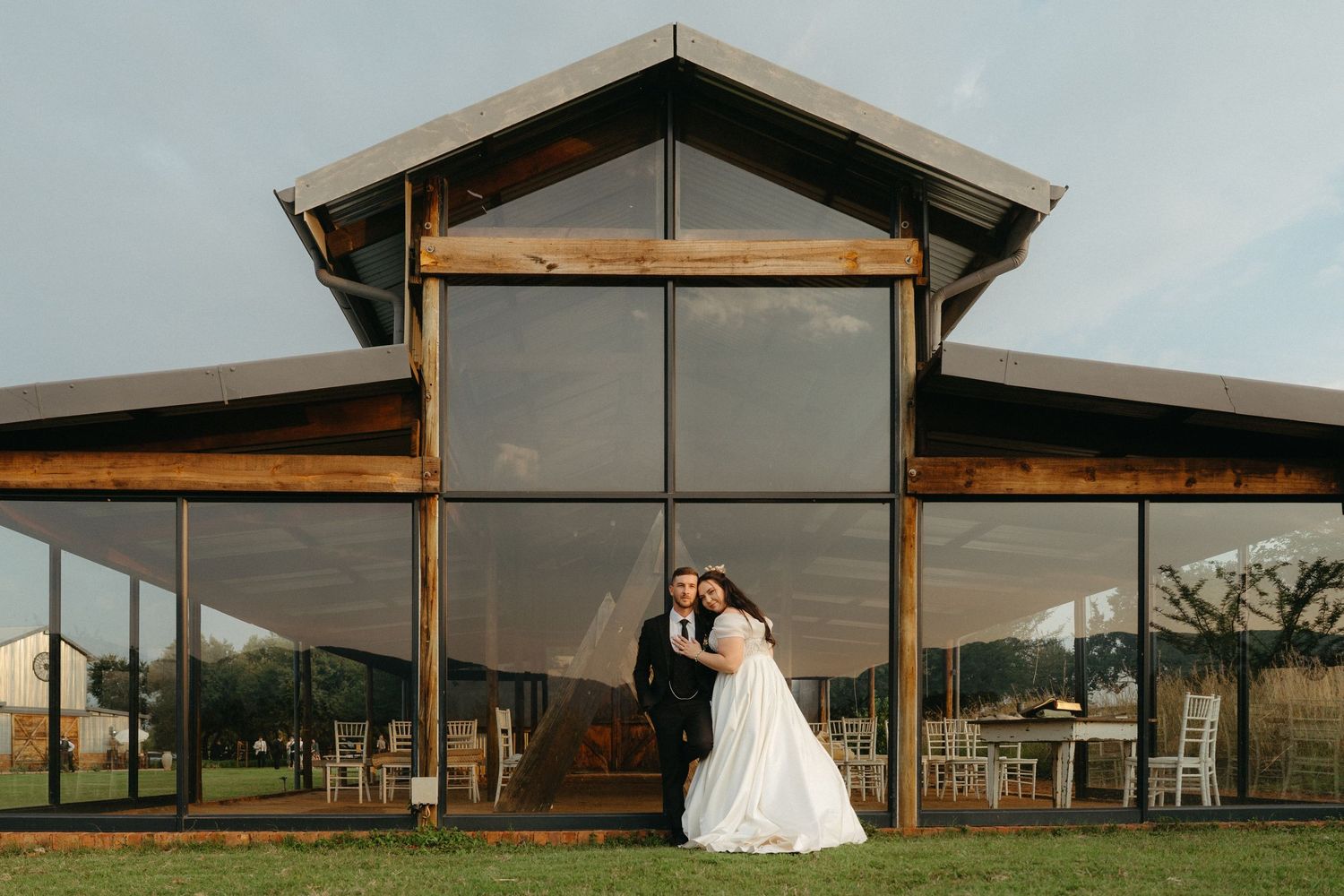 A wedding couple poses together under a modern rustic pavilion structure surrounded by scenic countryside views.