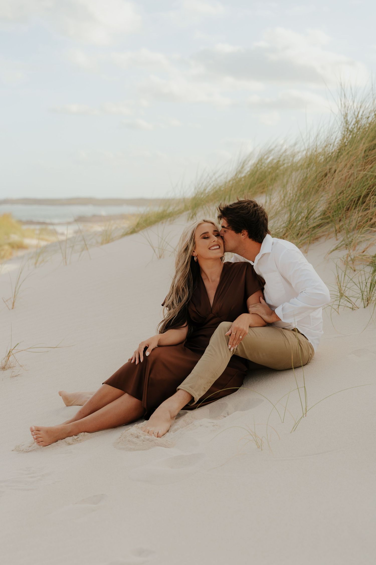 Couple sitting on sandy beach dunes, woman in brown dress smiling as man kisses her cheek.