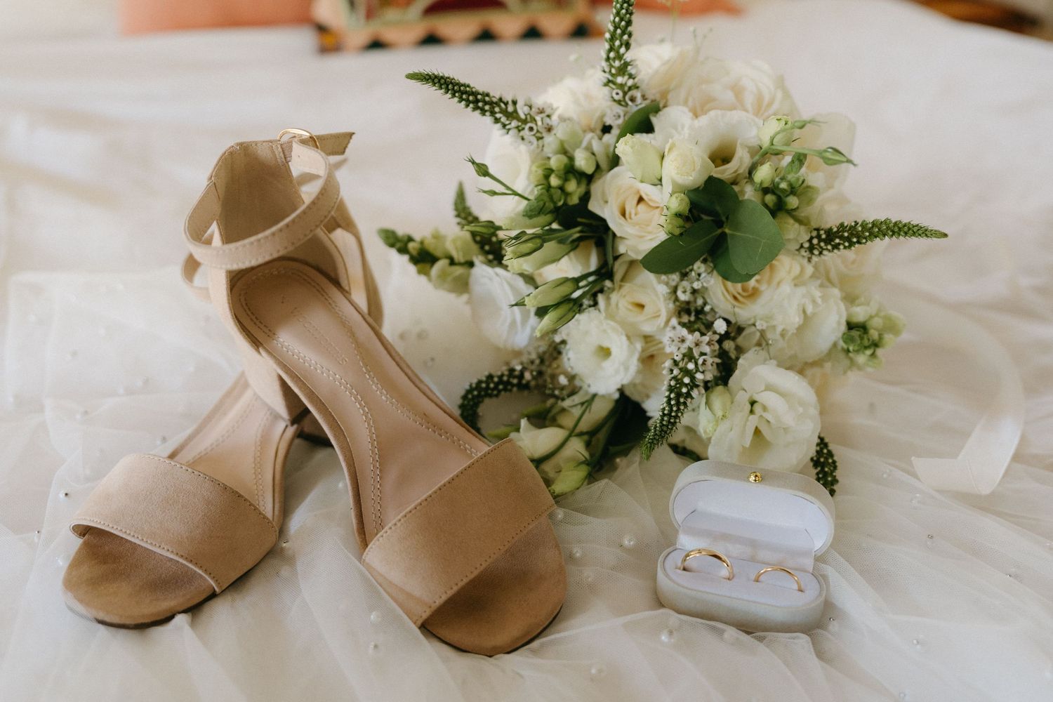 Elegant beige wedding shoes and a white floral bouquet with greenery arranged on white linen.