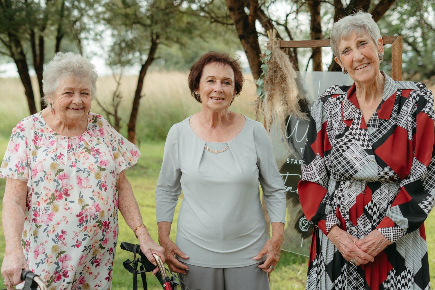 Three elderly friends standing together outdoors in casual clothing against a natural backdrop of trees and grass.