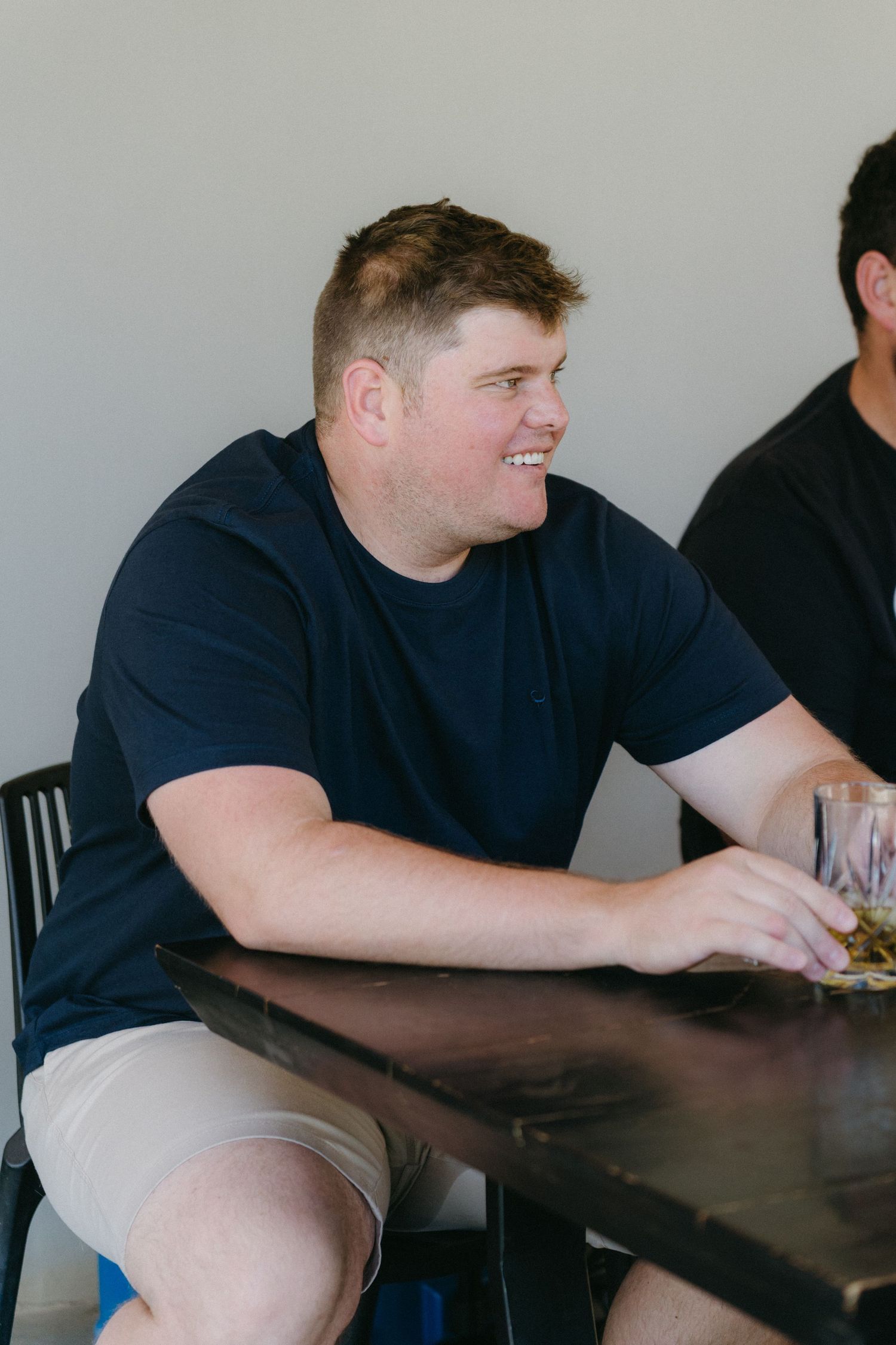 A person wearing a navy t-shirt sits at a dark table smiling during a casual gathering.