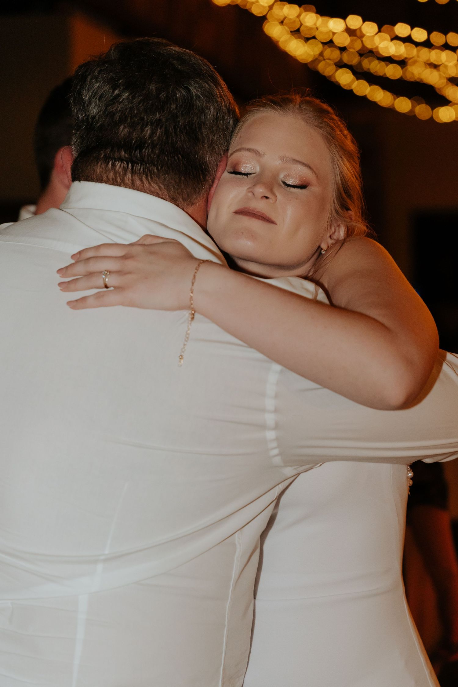 A romantic dance embrace under warm glowing string lights at a wedding reception.