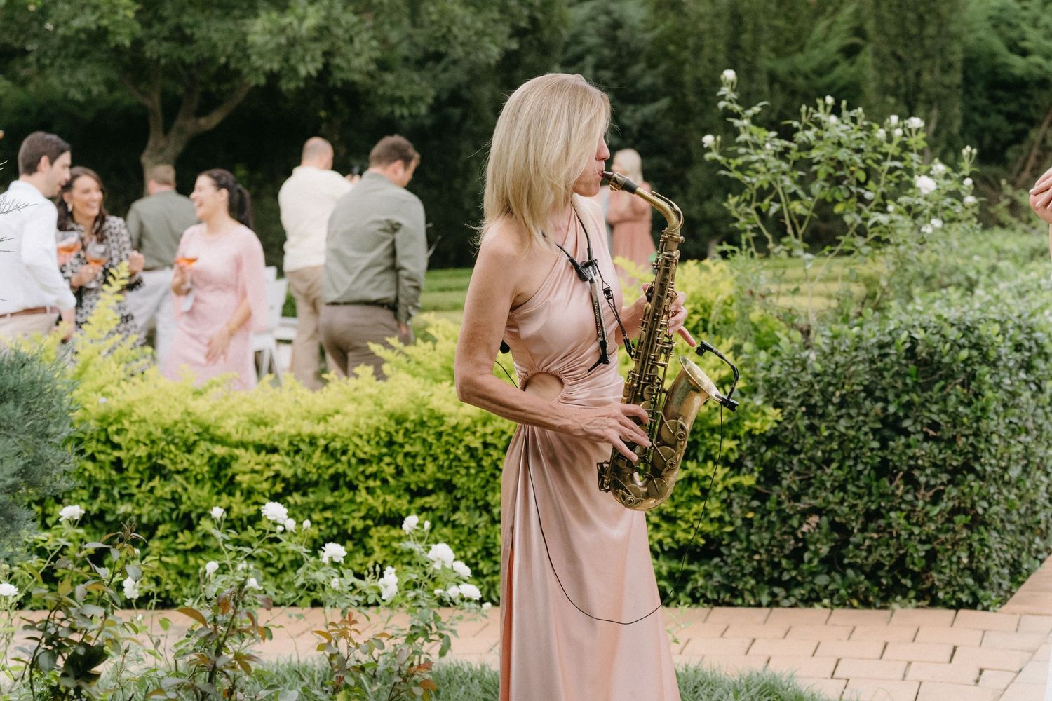A musician plays a saxophone in an outdoor garden setting with guests gathered in the background.