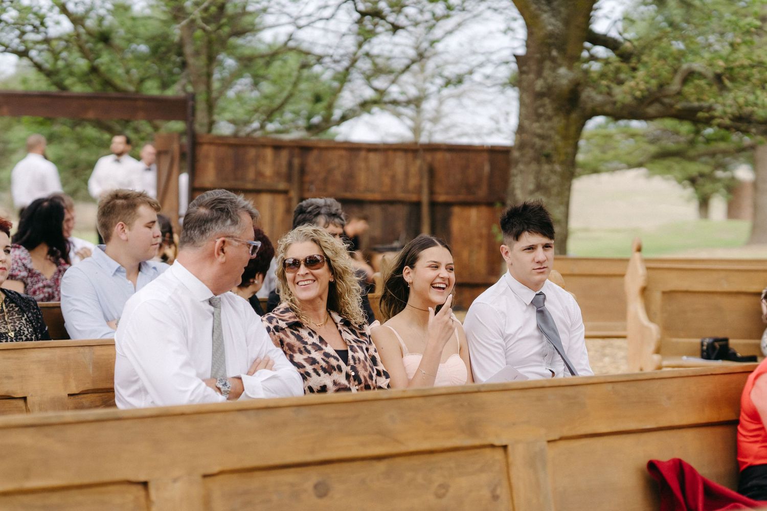 Wedding guests seated in wooden pews at an outdoor ceremony venue.
