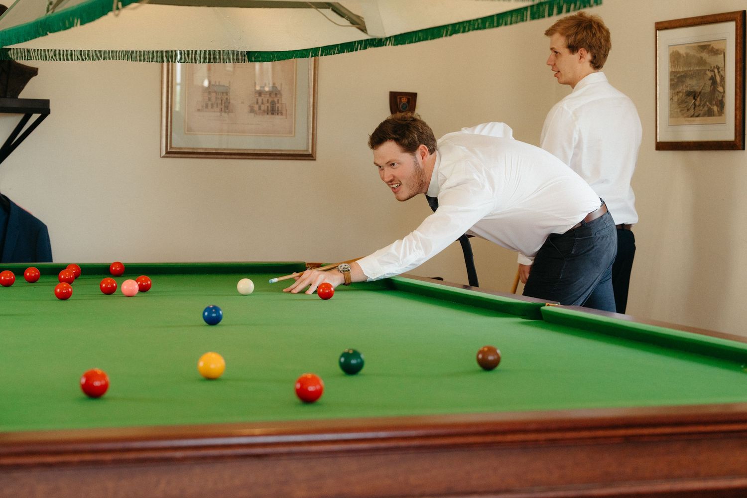 A groomsman lines up a shot on a green snooker table with colored balls scattered across the felt surface.