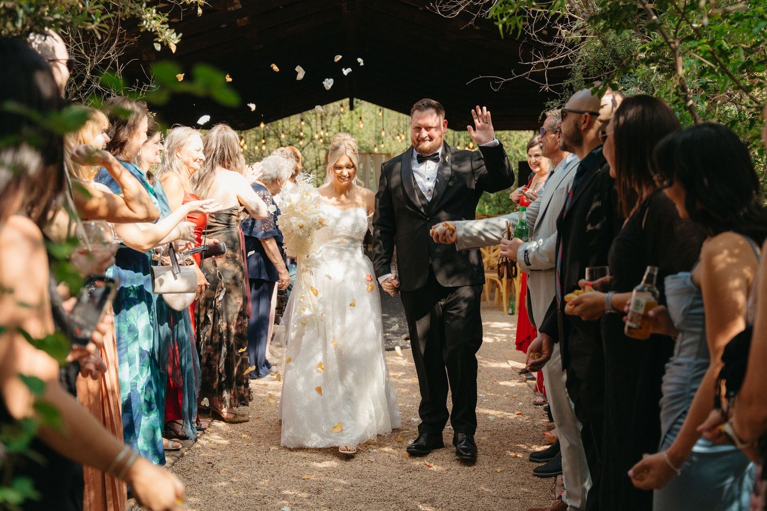 Wedding guests throw flower petals as newlyweds walk down outdoor path during recessional celebration.