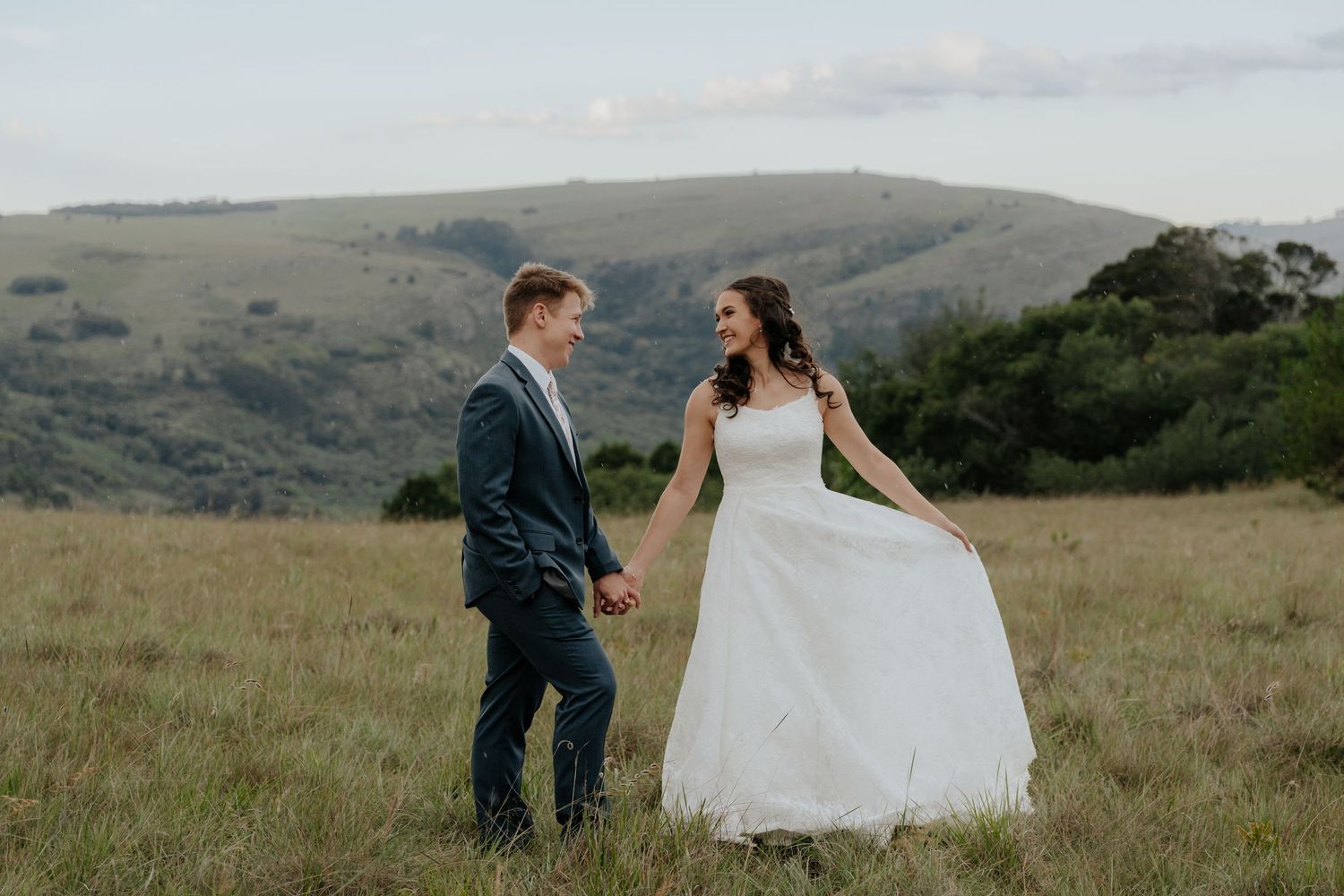 A couple in wedding attire stands together in a scenic mountain meadow with rolling hills in the background.