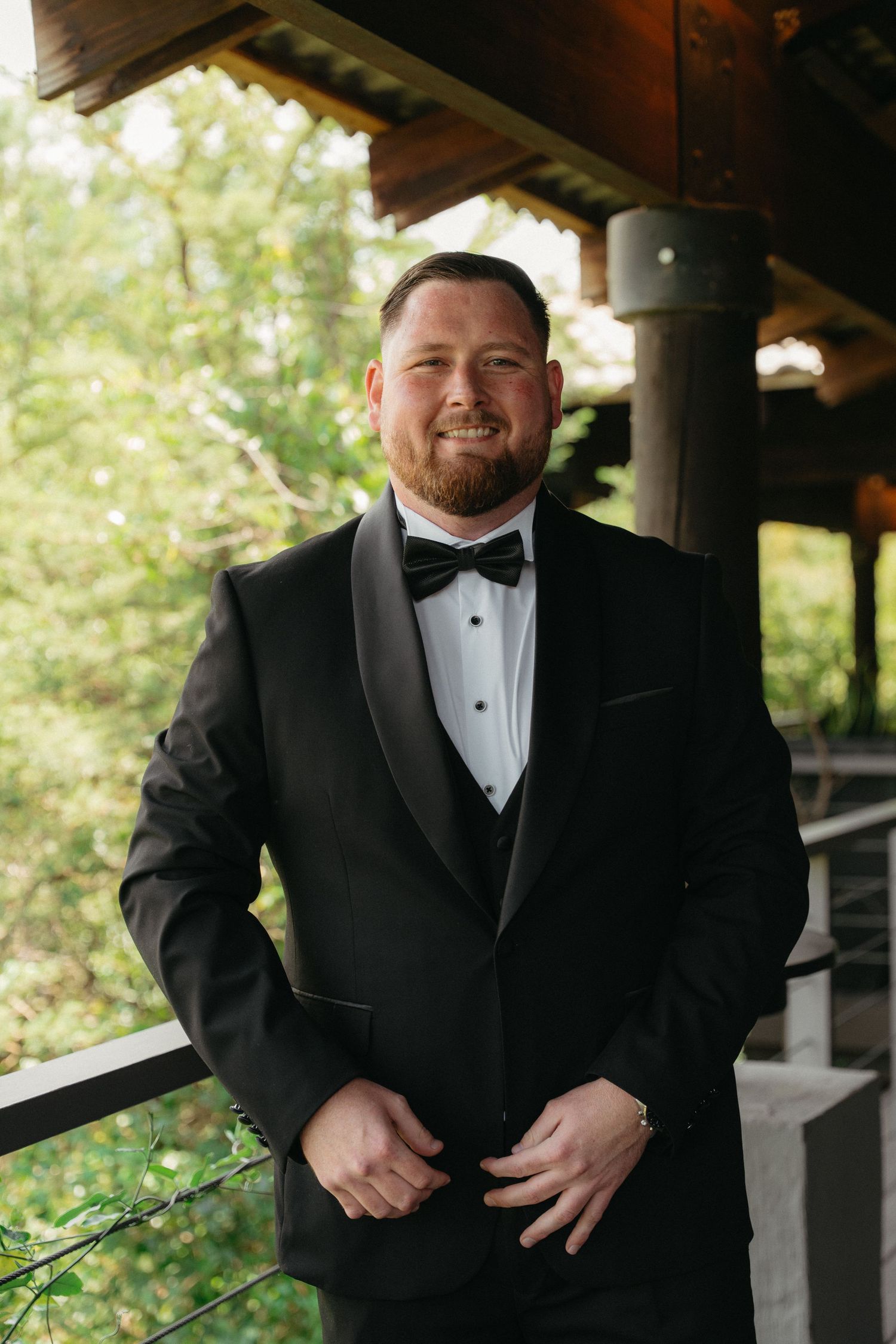 Groom in classic black tuxedo and bow tie poses on wooden porch with natural outdoor backdrop.