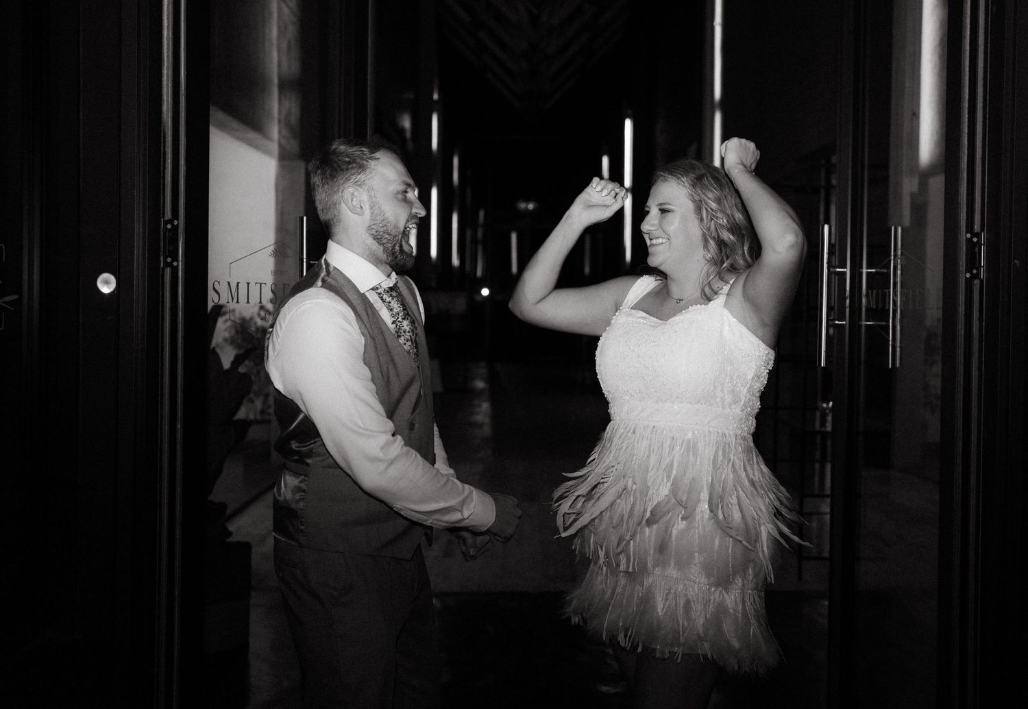A couple dances joyfully at their wedding reception, the bride wearing a flapper-style fringed dress under dim lighting.