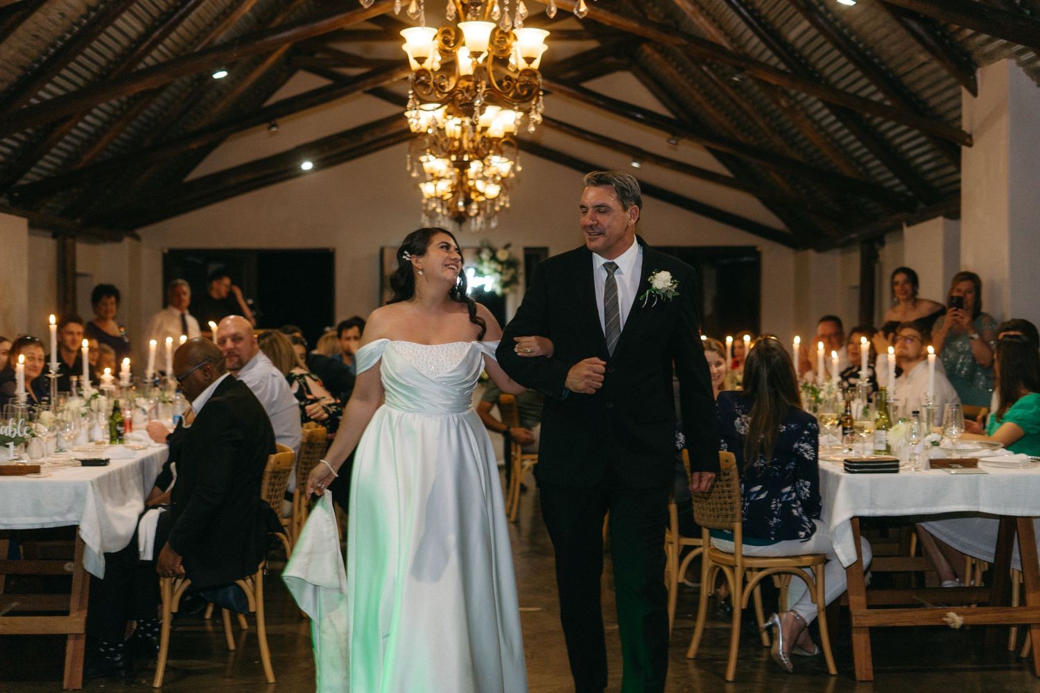 Elegant wedding reception in a rustic venue with exposed wooden beams, chandeliers and decorated dining tables.