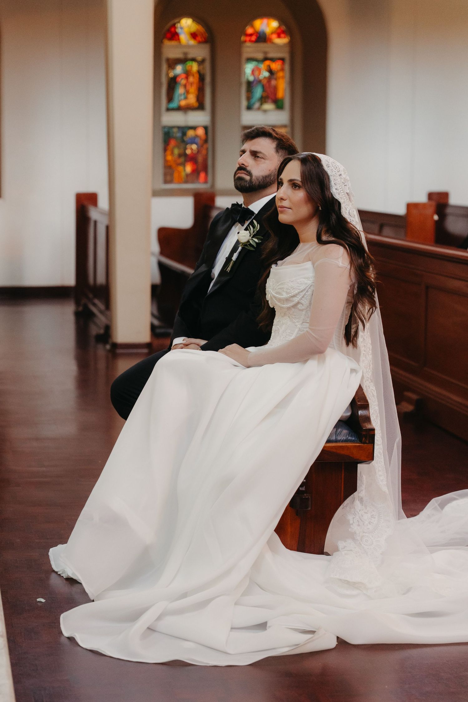 Bride and groom sitting together in a church pew, with stained glass windows in the background.