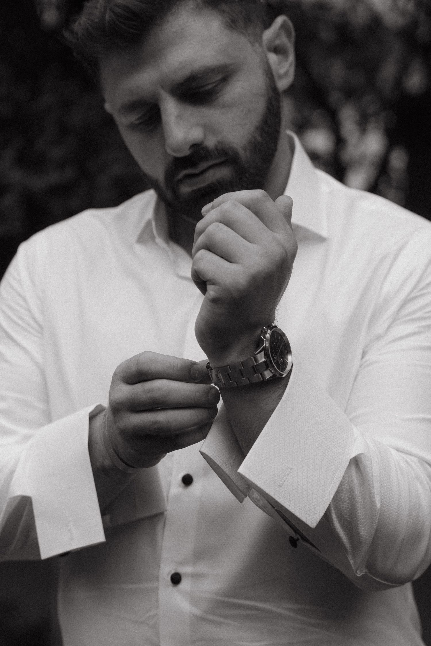 Black and white portrait of bearded man in white dress shirt fastening cufflinks while wearing a luxury wristwatch.