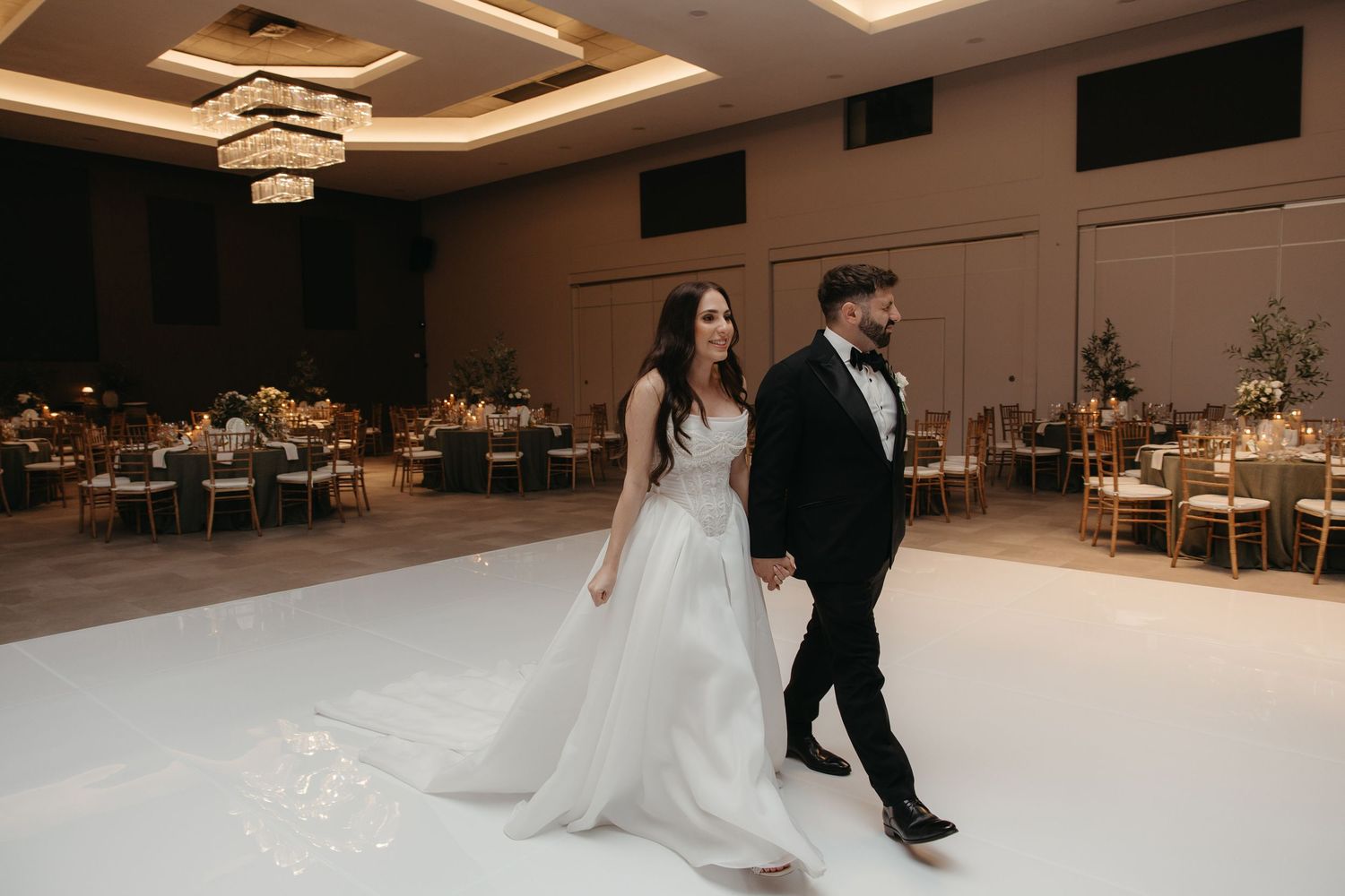 Bride in white gown and groom in tuxedo walk hand in hand on white dance floor in elegant ballroom.