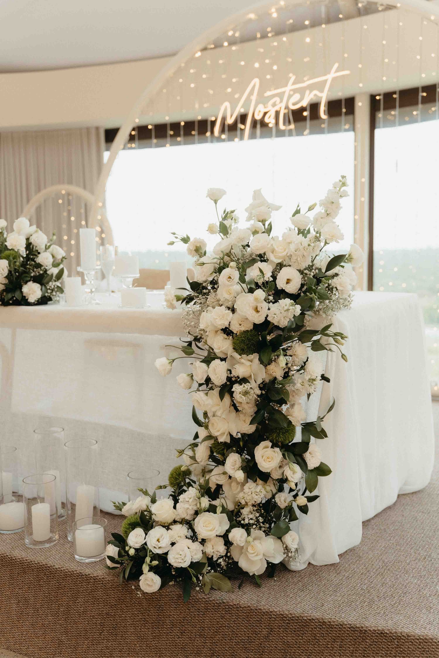 Elegant wedding sweetheart table adorned with lush white floral arrangements, greenery, and candles in a softly lit venue.