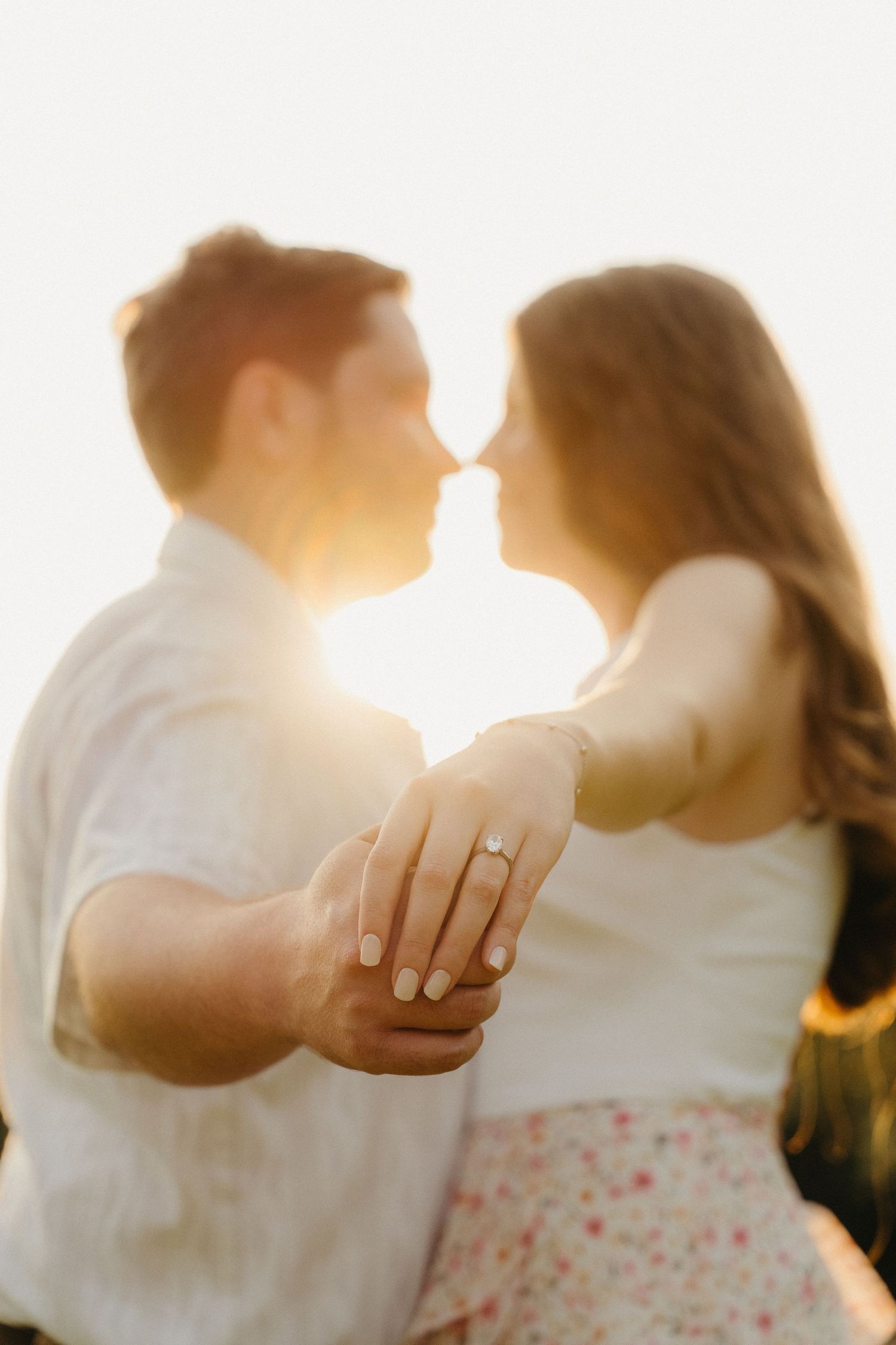 Couple sharing a romantic kiss at golden hour, woman displaying diamond engagement ring in the foreground.