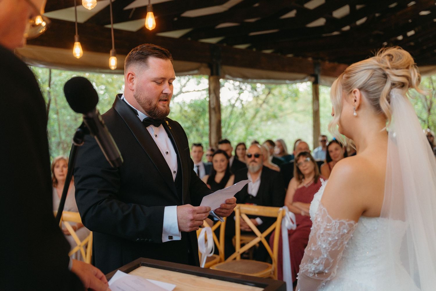 A wedding ceremony takes place under a covered outdoor venue with string lights and wooden chairs.