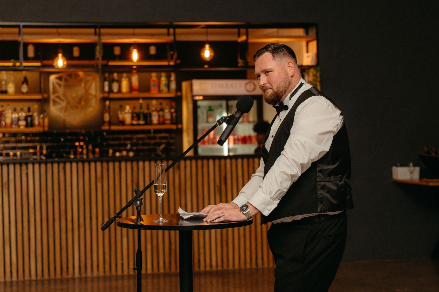 A performer in formal attire speaks into a microphone at a bar venue with warm ambient lighting and wooden decor.