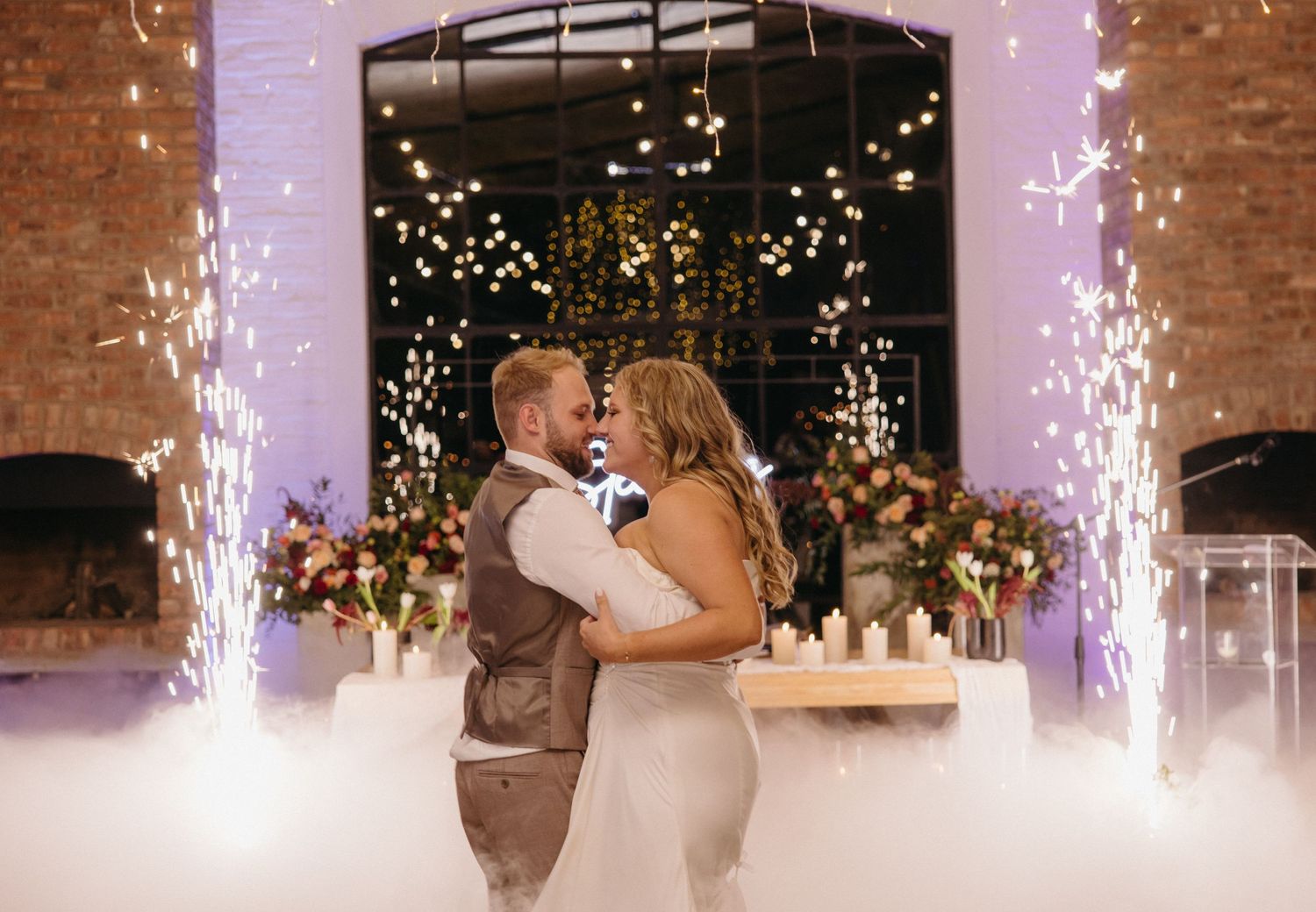 Couple shares a romantic dance moment with indoor sparkler fountains and twinkle lights at an elegant venue reception.
