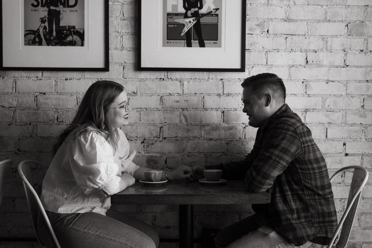 Two people sit across from each other at a small table with drinks in a cafe with exposed brick walls.
