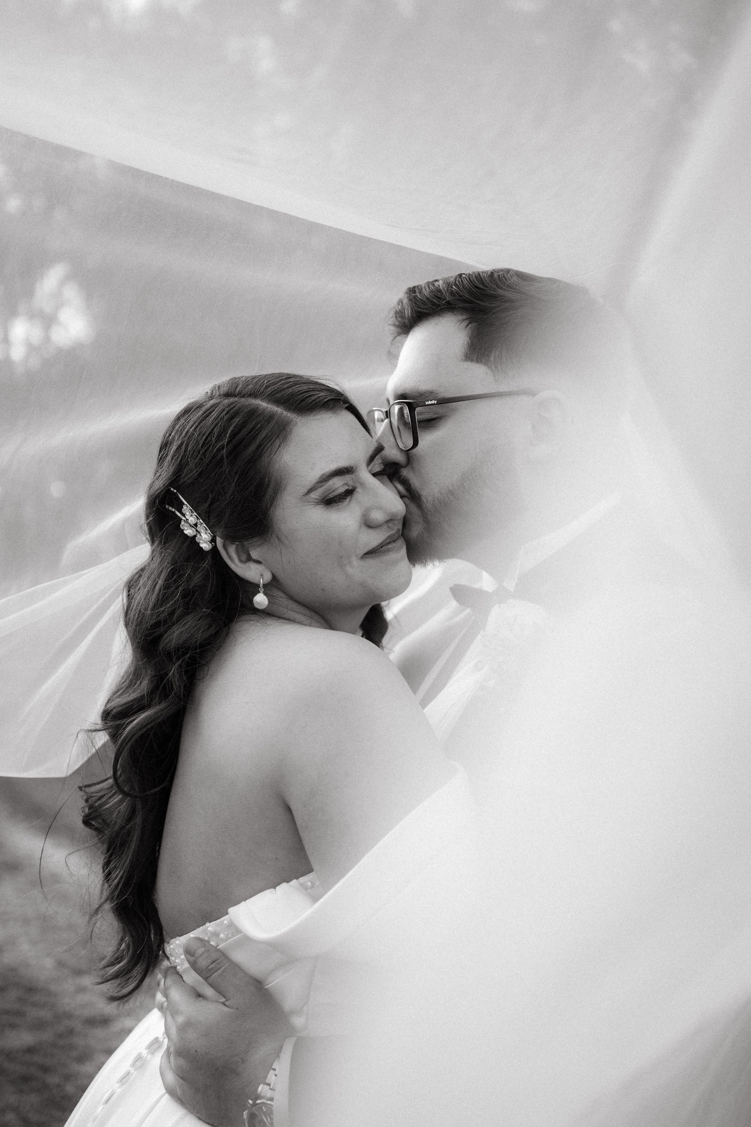 Black and white wedding portrait showing tender moment through sheer veil during romantic outdoor photoshoot.