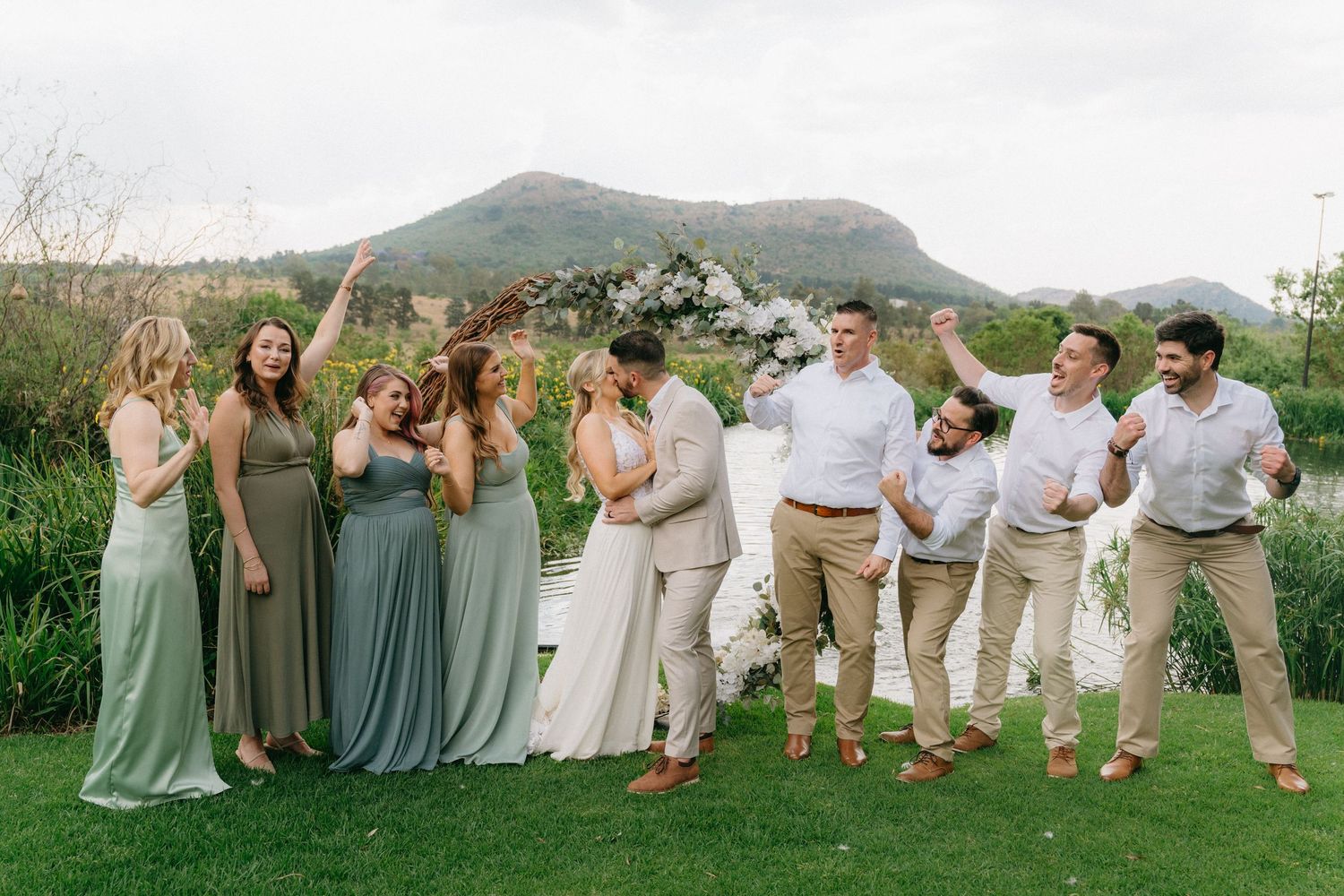 Wedding party members pose casually on grass with mountains in the background.