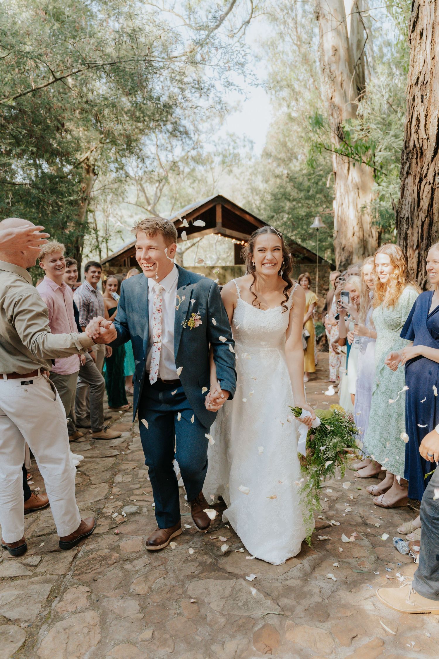 A joyful wedding celebration on a stone path surrounded by eucalyptus trees as guests shower the couple with confetti.