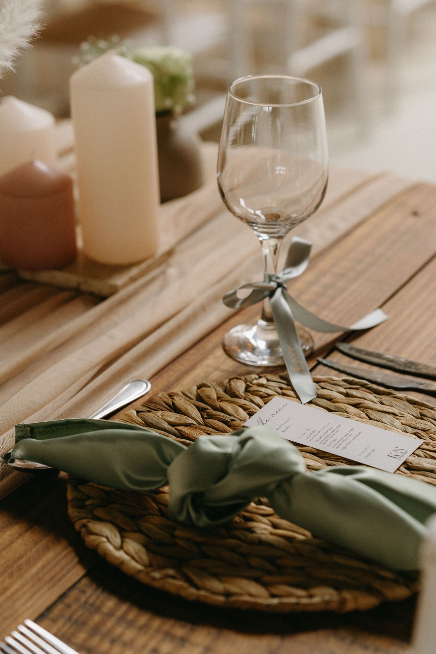 Elegant table setting with wine glass, white candle and sage green ribbon on woven placemat on wooden surface.