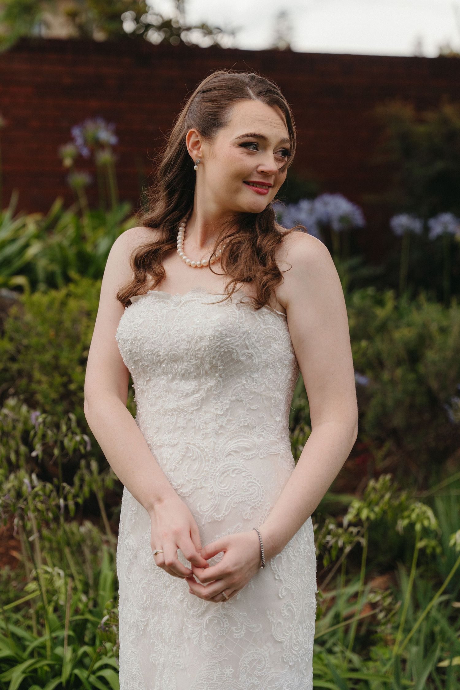 A bride in a white lace wedding dress poses outdoors in a garden setting with natural lighting and greenery.