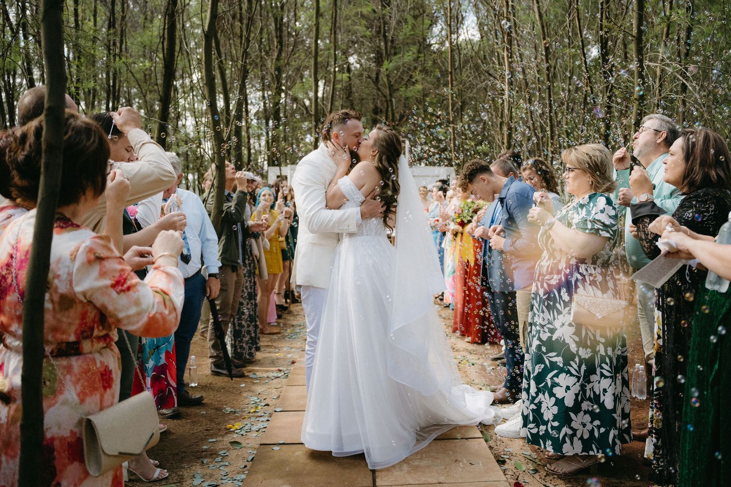 Wedding guests celebrate as the newlyweds share their first kiss on a rustic outdoor woodland path.