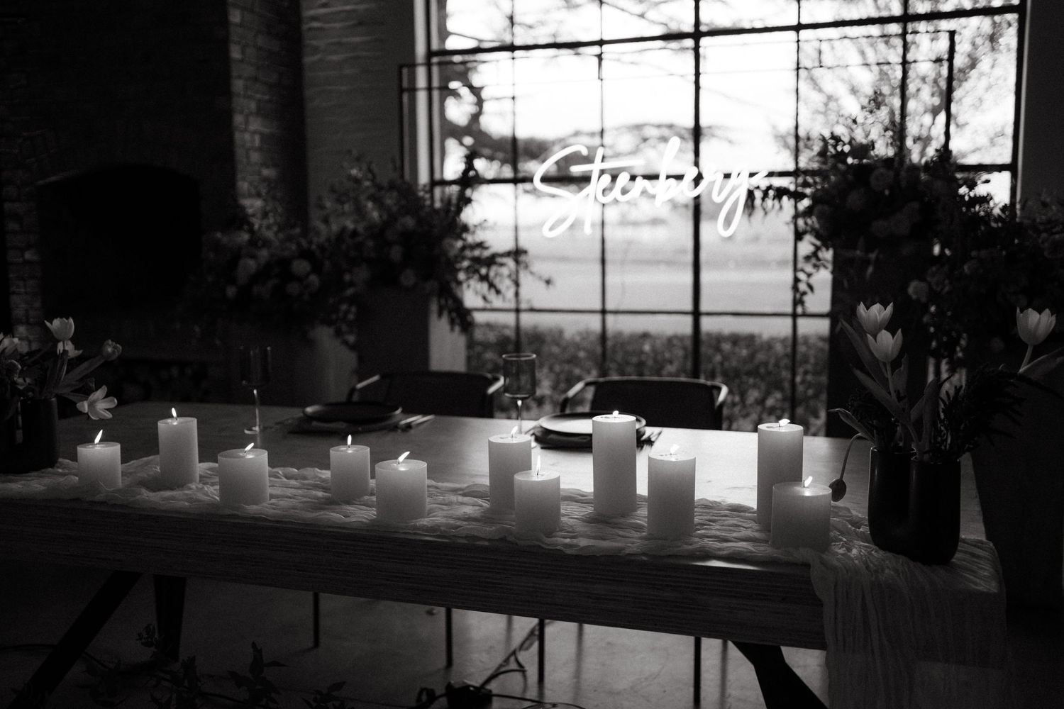 Black and white image of glowing candles arranged on a long table by a window.