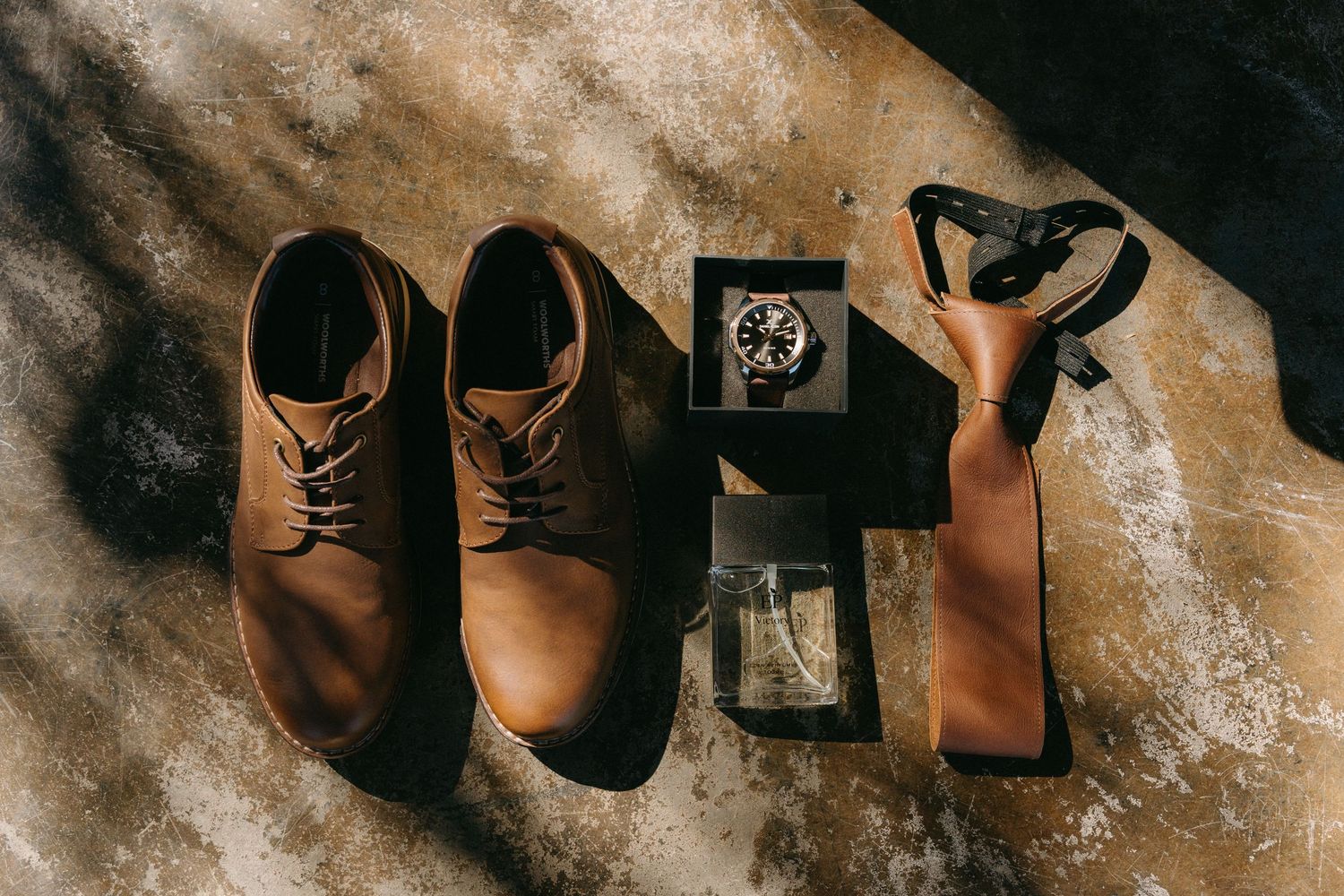 Wedding details flatlay featuring brown leather shoes, watch, and tie on textured surface.