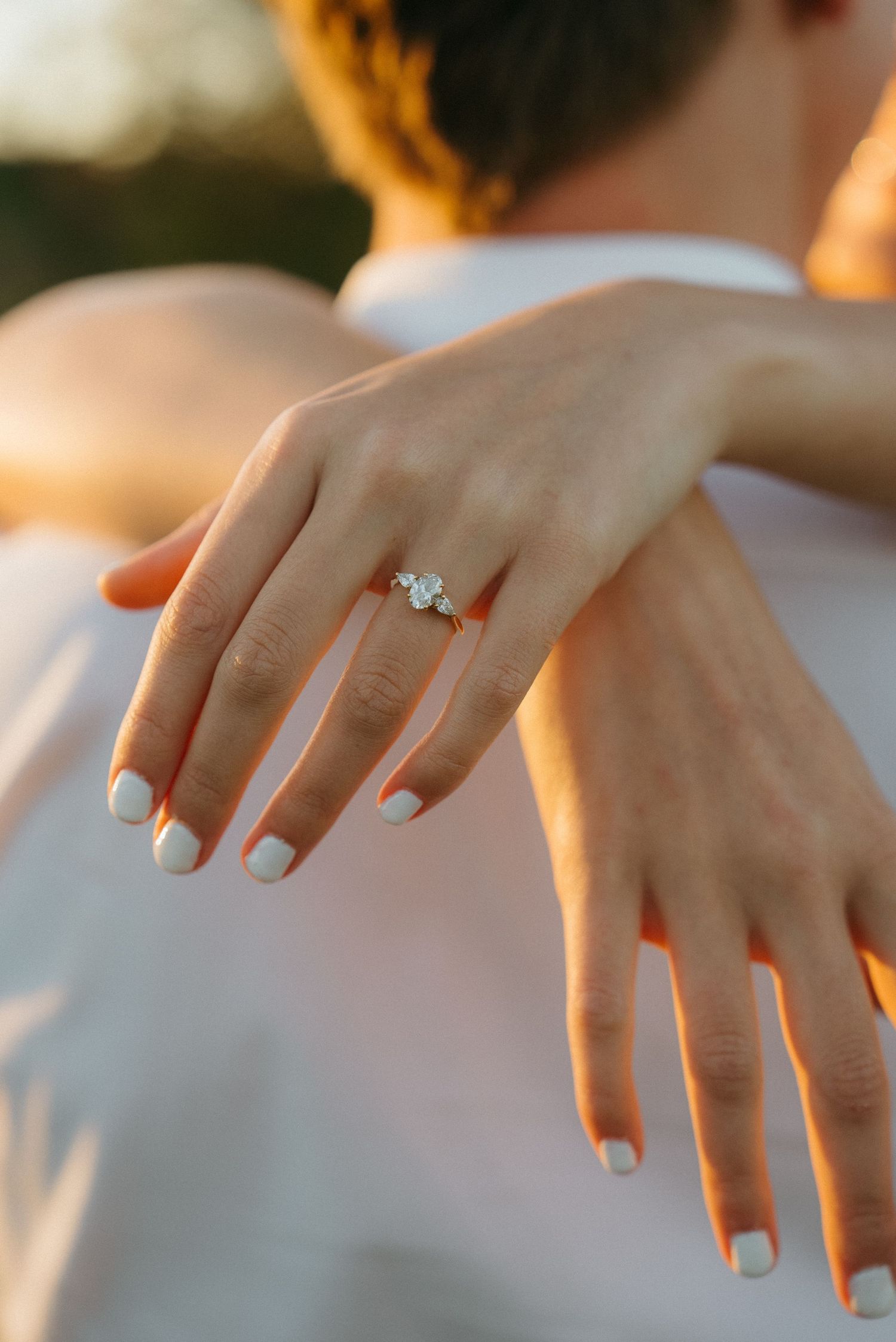 Close-up view of hands with French manicure displaying a solitaire diamond engagement ring in warm golden sunlight.