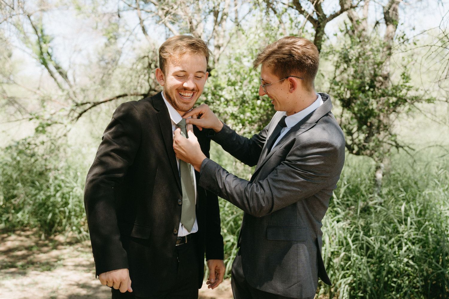 Two men in dark suits share a playful moment while adjusting ties outdoors.