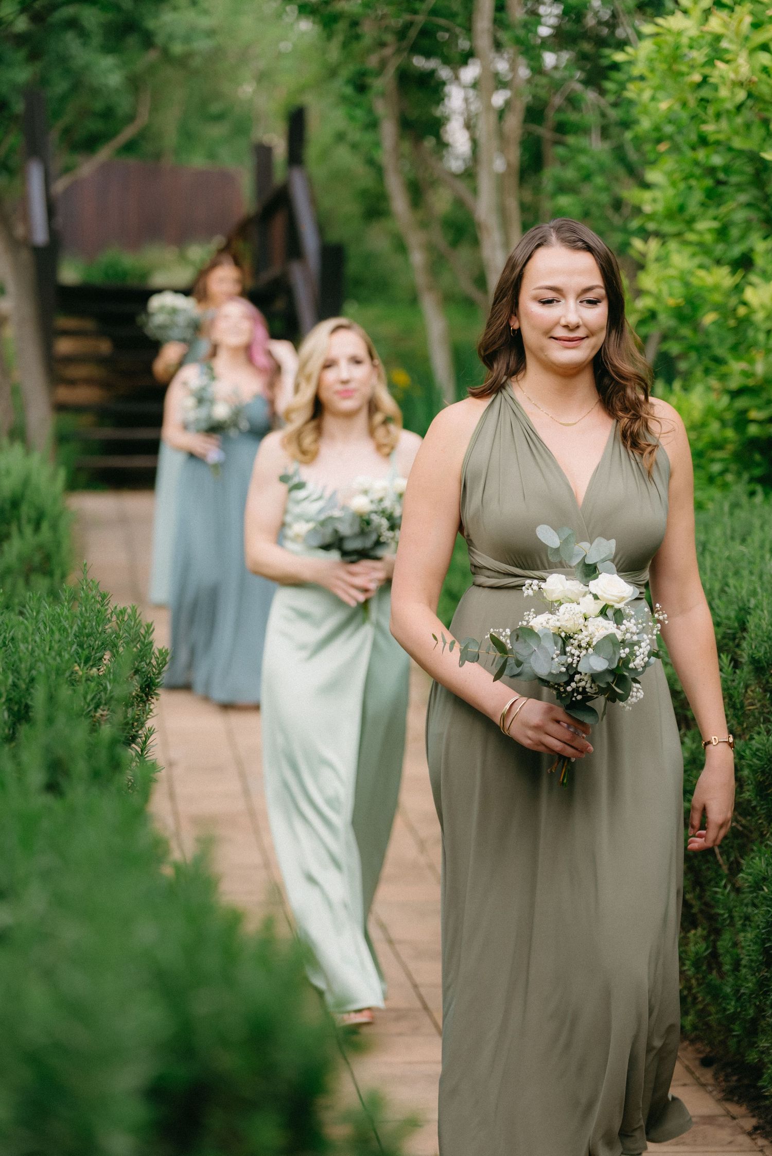 Bridesmaids in sage green dresses walk along a garden path carrying floral bouquets.